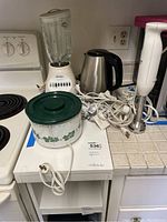 Overview of all kitchen appliances on a white cart and stove including Oster blender, crock pot, stainless electric kettle, handheld immersion blender, and tangled extension cords.