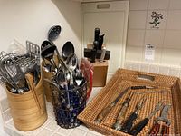 Wide view of various kitchen utensils in bamboo and metal holders, knife block in back, and woven tray holding decorative knives and utensils.