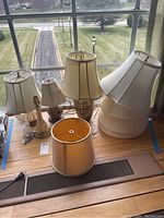 Four assorted table lamps and nine various lamp shades on a wooden surface by a window.