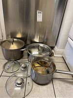 Three stainless steel pots of varying sizes with their matching stainless steel lids. Three extra glass lids with metal handles placed in front.