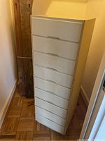 Tall narrow white chest of drawers with seven drawers, positioned upright against a wall on parquet flooring.