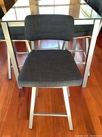 Front view of one swivel bar chair showing dark blue upholstery, metal frame and footrest with table legs and glass top visible behind