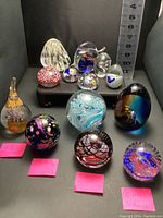 Front view of all 13 paperweights shown on black surface with measure behind for scale. Colorful and clear designs with labels visible.