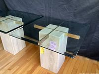 View of two travertine block side tables with clear glass tops supported by brass crossbars on hardwood floor.