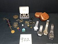 Full assortment of men's jewelry items arranged on black background including wristwatches, cufflinks, lapel pins, money clips, tie clip, and leather pouches.