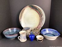 Group photo showing 8 assorted pottery items including plates, bowls, mug, creamer, teapot, blue vase and small cup arranged on a black background.