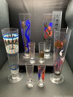 Full view of 8 glasses including 4 tall beer flutes and 4 smaller aperitif glasses, showing colorful decals and artist names on bases.