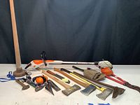 Full view of the lot including Stihl electric weed eater, axes, entrenching tool, ear protection and more spread out on a white surface with black background.
