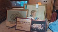 Front view of multiple framed artworks including sampler, paintings, prints, leaning against furniture