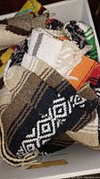 Box containing multiple folded woven blankets with varied geometric patterns and colors including black, white, brown, orange, green, and fringed edges.