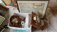 Photo showing two brass ship's clocks mounted on wood plaques inside a cardboard box with bubble wrap, and a framed nautical map leaning in the background.