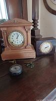 Three clocks on a wooden surface including a Seth Thomas mantle clock with key, a GE mantle clock, and a small round quartz clock.