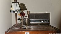 Photo of three lamps including cranberry glass lamp, brass blow torch lamp, and stained glass mosaic shade lamp, along with vintage Magnavox radio on wooden surface.