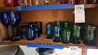 Shelf displaying blue tall-stem wine glasses, blue short-stem textured glasses, blue stemless textured glasses, and green stem and stemless glasses in different sizes.