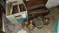 Photo showing wooden box of hand tools, cardboard box of tools, brass circular parts, and brass rectangular box.