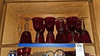 Seven red handblown glass stemware pieces including six goblets and one smaller glass with clear stems and bases, arranged on a wood shelf.