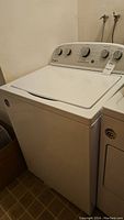 Front view of white Whirlpool top-loading washing machine in basement showing closed lid and control panel with multiple dials.
