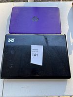 Two HP laptops lying closed on a white surface, showing the top covers. The purple laptop has a matte texture with an HP emblem, the black laptop has visible scuffs and an HP logo.