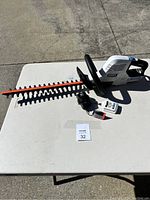 Photo of white and black HART 20V Hybrid battery powered hedge trimmer with an orange blade cover alongside a black charger and a white charger base with indicators arranged on a white table outdoors.