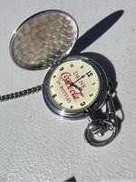 Photo showing the open pocket watch face with Coca-Cola branding and chain shadow.