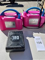 Two pink electric balloon pumps with blue handles and attached nozzles shown on a surface with power cords visible, along with a black power adapter labeled with voltage and capacity.