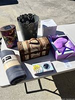 Wide shot of the entire lot showing all items on a white table including decorative chest, metal container, blanket, gift boxed eye mask and pillow, cylindrical butterfly container, aromatherapy timer, and an additional white box.