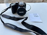 Front view of Kodak PIXPRO AZ421 with lens cap on and strap extended on table, showing camera body and lens cap with Kodak logo.