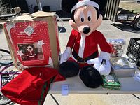 Photo of the large animatronic Mickey Mouse figure in Santa outfit with its original box, a red fabric bag, and Christmas ornament balls on a table.
