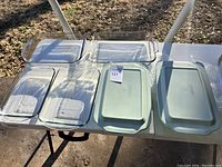 Clear glass dishes and plastic lids arranged on white folding table outdoors with natural light.