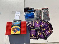 Red Lego baseplate, small blue Lego bricks, several purple Pokémon Trick or Trade trading card packs, and two black and red Allure trading card packs displayed on a white surface.