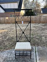 Full view of black metal hall tree standing outdoors on textured concrete ground, showing cushioned bench seat at bottom and hooks along vertical back frame.