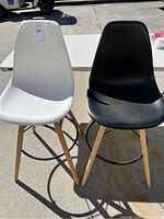 Pair of modern barstools, one white and one black molded plastic seats, wooden legs, and black metal footrest rings, photographed outdoors on cement pavement with a white table and a vehicle in the background.