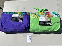 Two folded outdoor blankets, one purple and one green with floral print, placed side by side on a table.