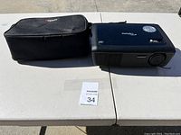 Black Optoma projector alongside a black carrying case on white table surface under sunlight.