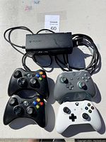 Top view of the black Xbox 360 power supply adapter with cables and four Xbox controllers: two black Xbox 360 wired controllers, one grey Xbox Series X/S wireless controller, and one white Xbox Series X/S wireless controller.