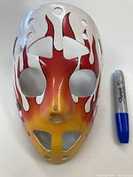 Front view of the 1970s fibreglass goalie mask with red, white, and yellow flames graphic, next to a blue Sharpie marker for scale.