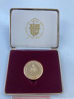 Medal displayed inside an open red velvet-lined box with cream lid showing gold event logo and text.
