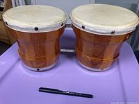 Pair of connected wooden bongo drums with natural hide heads and metal tacks, showing external wear consistent with age.