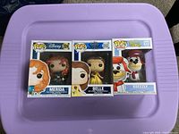 Three Funko Pop boxed figures on a purple surface: Merida, Belle, Breezly.