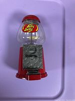 Front view of red and clear plastic Jelly Belly candy dispenser showing coin slot and dispenser opening, with Jelly Belly logo on clear container.