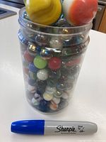 Clear plastic jar filled with a variety of colorful marbles including some oversized yellow and orange marbles on top, with a blue Sharpie for scale.