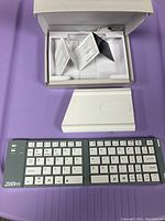 Zoom Bluetooth keyboard with protective case and user manual laid out on purple surface. Keyboard is compact with white keys and grey frame, 'Zoom' branding visible.