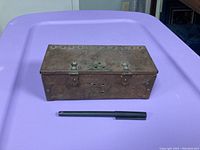 Top view of a rusted rectangular metal box with rivet accents and two latches, placed on a purple surface next to a black pen for scale.