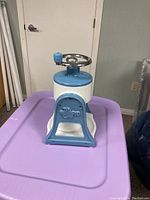 Front angle view of the Pampered Chef ice shaver showing the blue and white body and chrome handle on a purple surface.