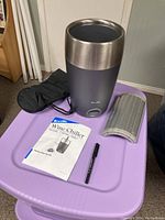 Wine chiller with soft carrying case, cooling element, manual, and pen on a purple table.