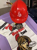 Red Ottawa Senators hardhat, hockey puck partially under hardhat, and one Official Woo-Hoo Towel laid out
