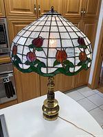 Full view of the lamp on a white surface with the Tiffany style stained glass shade lit, showing red and orange flowers, green leaves, and grid pattern on white glass.