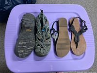 Top view of two pairs of women's sandals on a purple surface. One pair is bulkier gray and teal Keen outdoor sandals and the other is sleeker black thong-style sandals.