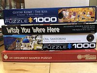 Photo of four jigsaw puzzle boxes stacked, showing names and piece counts.