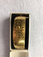 Close-up top view of the gold plated bangle showing detailed engraving and attached safety chain inside a display box.